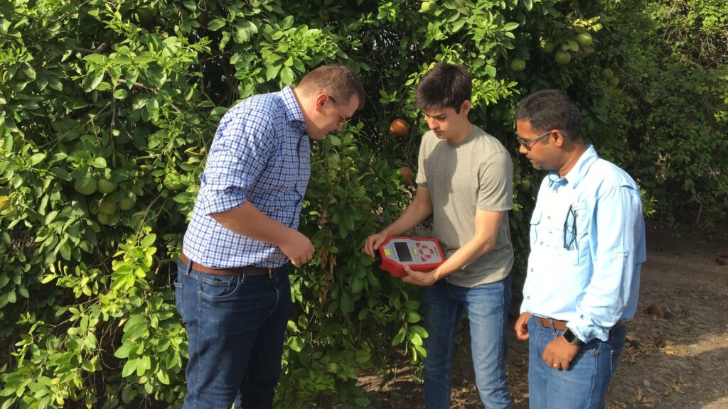 Two students in an orange field operate a handheld digital device as a professor standing to the right of the students supervises their work.