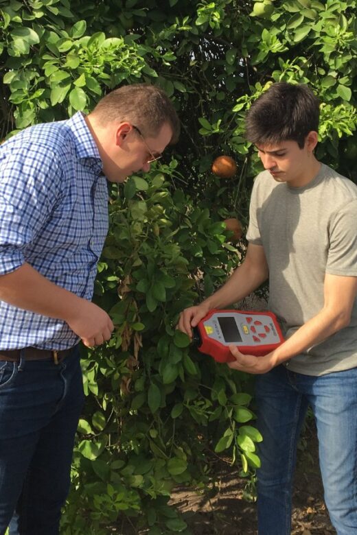 Two students in an orange field operate a handheld digital device as a professor standing to the right of the students supervises their work.