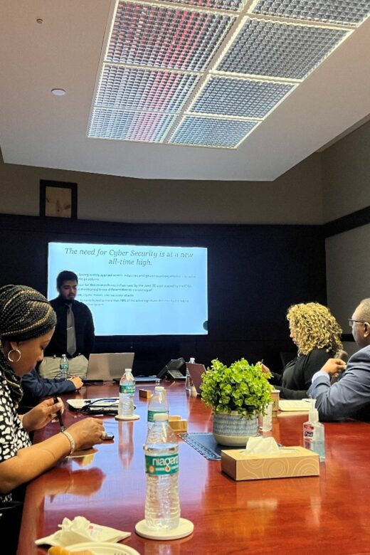 A student stands at a podium in front of a white video screen with a wood conference table of five people in professional attire